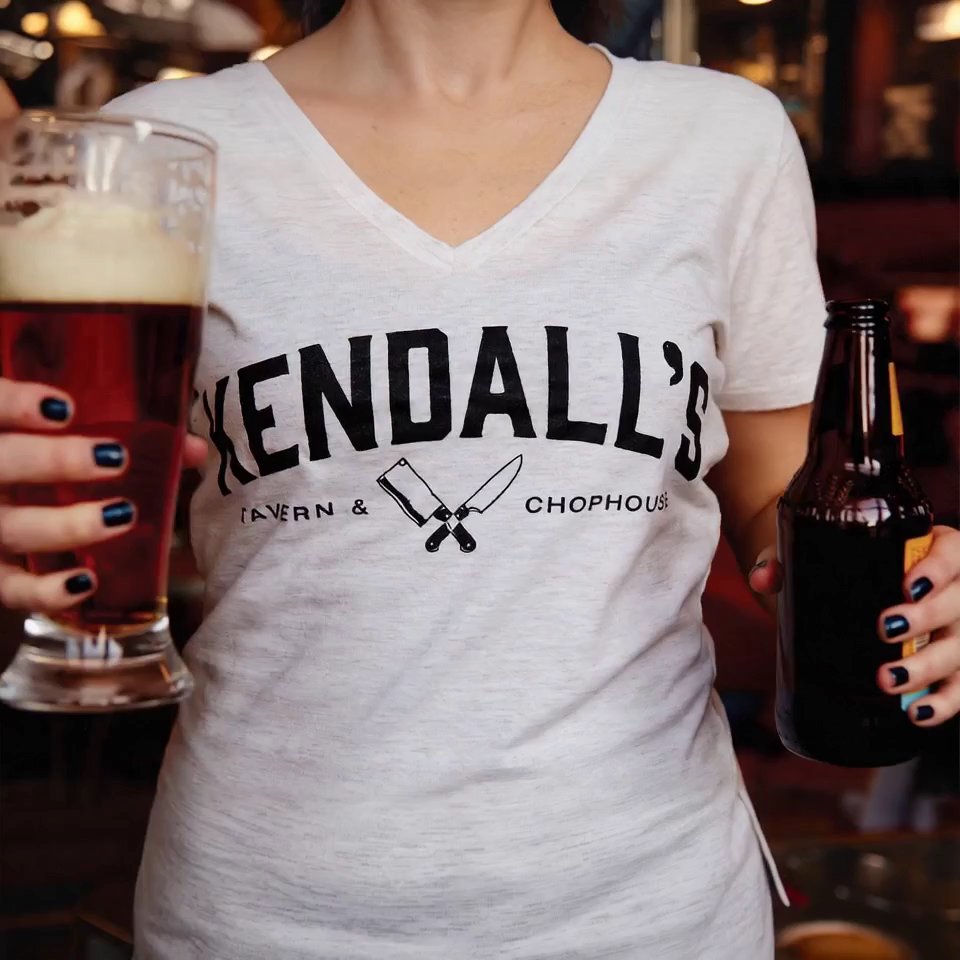 A woman in a Kendall’s Tavern & Chophouse T-shirt holding a pint of beer and a bottle